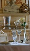Mit ziselierten Gläsern, Kieselsteinen und weisser Rose in hellen Farben gedeckter Tisch; im Hintergrund antike Anrichte aus Naturholz
