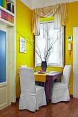 Bold yellow in corner of living room - tiny seating area with white covered chairs at wooden table below window