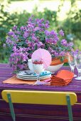 Preparations for a summery garden table set with bougainvillea on a purple tablecloth and back of chair in the complementary colour of yellow