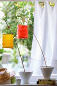 Small paper lanterns attached to wooden canes with clothes pegs in garden setting with white curtain in background