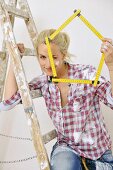 Renovating - woman sitting on ladder holding up a folding ruler