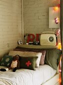 Knitted doll and stack of scatter cushions against headboard of bed below retro radio on shelf against whitewashed brick wall
