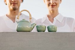 Teapot and tea cups sitting on ledge, cropped view of man and woman waiting nearby