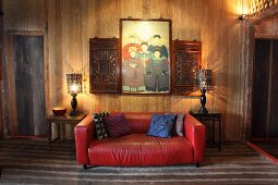 Red leather couch flanked by two side tables against plain wooden wall and below family portrait and carved wooden cabinets