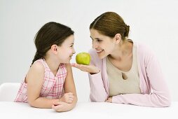 Mother and daughter sitting face to face, smiling at each other, woman holding apple