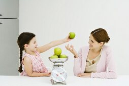 Mother and daughter weighing apples on scale, smiling at each other, girl holding up one apple