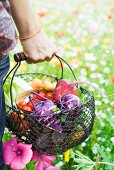 Person standing in field of wildflowers, carrying basket full of fresh produce, cropped view