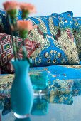 Turquoise vase of flowers on glass table in front of couch with Oriental upholstery and scatter cushions