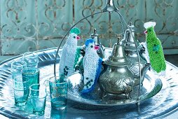 A round metal dish holding Oriental teapots with knitted finger protectors on the handles, with coloured tea glasses