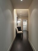 Upholstered chair next to chest of drawers in narrow corridor with recessed spotlights