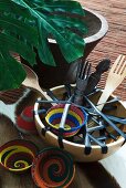 Schale mit Salatbesteck aus Holz und farbigen Schälchen in afrikanischem Stil neben Topfpflanze auf Boden