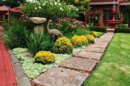 Red fountain in well-tended, flowering garden with stone-flagged path and statue of bird