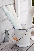 Rolls of paper in enamel bucket and tealight holder shaped like a miniature lighthouse on teak terrace; painted weatherboard cladding in background
