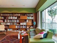 Designer sofa in front of a bank of windows and wooden book shelves on a green wall in a modern living room