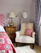 Upholstered armchair and vintage standard lamp next to painted bedside cabinet against pastel wall in corner of bedroom