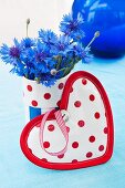 Heart-shaped pot holder in front of fabric-covered beaker of cornflowers