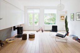 Minimalist living room with black armchairs on wooden floor and pouffes in front of small, round, iron stove in front of terrace doors with view of garden