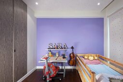 Bed in front of a window with a fabric roller blind and writing desk in front of a lavender wall in a small child's bedroom