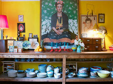 Large self-portrait of Frida Kahlo above middle of open-fronted shelves of crockery surrounded by various pictures and painted china against yellow wall