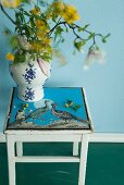 Blumenvase aus Porzellan mit floralem Muster auf einem Holzhocker mit aufgemaltem Vogelbild