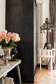 Silver, trophy-shaped vase of roses on wooden bench next to roller-door cupboard and iron lamp on side table