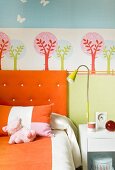 Farbe im Kinderzimmer - Bettwäsche und Kopfpolster in Orange vor Wandmalerei in Schablonentechnik mit stilisierten Bäumen