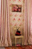 Unusual hat with feathers and red hatband on antique chair against wall with floral wallpaper and striped, floor-length curtains at windows