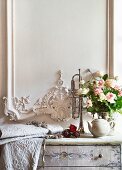 Bouquet of roses and embroidered pale fabric on chest of drawers delicately painted in soft colours below white wooden wall panel with ornate carved details in elegant, country-house style