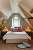 Stacked scatter cushions on double bed below window in painted gable end of attic storey, romantic bedside lamps and round cushions on floor
