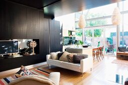 Pale, modern sofa next to black living room cupboards and view into open-plan kitchen-dining room in front of glass facade