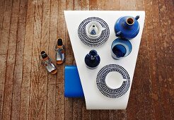 Trapezoidal shaped white table with vintage cutlery and blue vases and old wood plank flooring; next to it, silver sandals