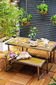 Holzgefliester Innenhof mit Esstisch und Sitzbänken; im Hintergrund ein weisser Kasten mit Bambuspflanzen und hängenden Pflanzentöpfen