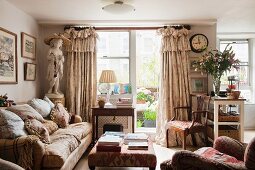 Englisches Wohnzimmer mit Blick auf den Balkon; üppige Volantvorhänge, viele Kissen auf dem Polstersofa und eine Statue in Mädchengestalt, vermitteln ein nostalgisches Flair