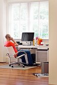 Simple office with flat screen monitor below window with blinds; man speaking on phone sitting in comfortable office chair at desk