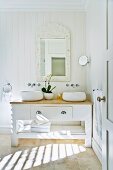 Solider Waschtisch aus Holz im Landhausstil mit darüberhängendem, gerahmten Spiegel und runden Waschschüsseln; dazwischen weiße Orchideen