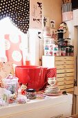 Oversized red teacup, tea service and Oriental doll on surface with sewing utensils in storage jars in background on chest of shallow drawers