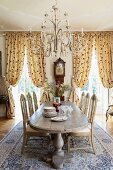 Magnificent dining room with solid wooden dining table below pretty chandelier; gathered, floor-length curtains in background at windows with view of park