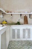 Corner of Moroccan kitchen with pale tiled worksurface, white base units with perforated wooden doors, floral grey and white stone floor and curved brackets supporting crockery shelf on two walls
