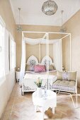 Oriental four-poster bed with painted headboard in bright, simple bedroom with mosaic stone floor and upholstered metal chair next to white side table