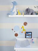 Nursery wall decorated with pale blue stripes and circles; various soft toys on floating shelf and pale blue changing mat on white changing unit