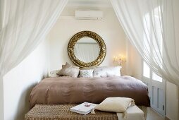 Stately bed behind open curtains; large, round, gilt-framed mirror above head