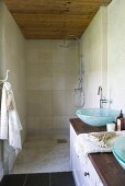 Shower head in narrow shower room with wooden ceiling; long wooden washstand with two glass washbasins in foreground