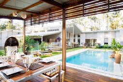 Dining table on roofed terrace with view of pool and luxurious, one-storey white house with pitch roof