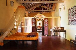 Schlichtes Doppelbett mit Baldachin im gelb getönten Schlafzimmer und Blick durch offene Türen in geschnitzten Holztrennwänden