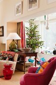 Decorated Christmas tree and wrapped presents in bay window; deep pink armchair and small, red, glass table shaped like demijohn in foreground