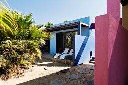 Contemporary sun loungers on the terrace of a blue hotel building looking out to a courtyard with round paving stones and large Mexican Fan Palm plant