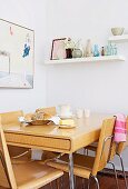 Dining area with pale, laminated timber furniture and collection of glass bottles on floating shelf in kitchen