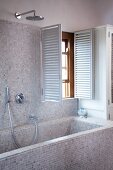 Bathtub and wall with bright, mosaic tiles and open shutters at the window in a modern bathroom