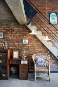 Vintage bureau, small cabinet and wicker chair in front of staircase against brick wall in loft-style interior