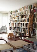 Large bookcase with white shelves, wooden sculpture leaning on bookcase, books on table in foreground, modern reading chair in background